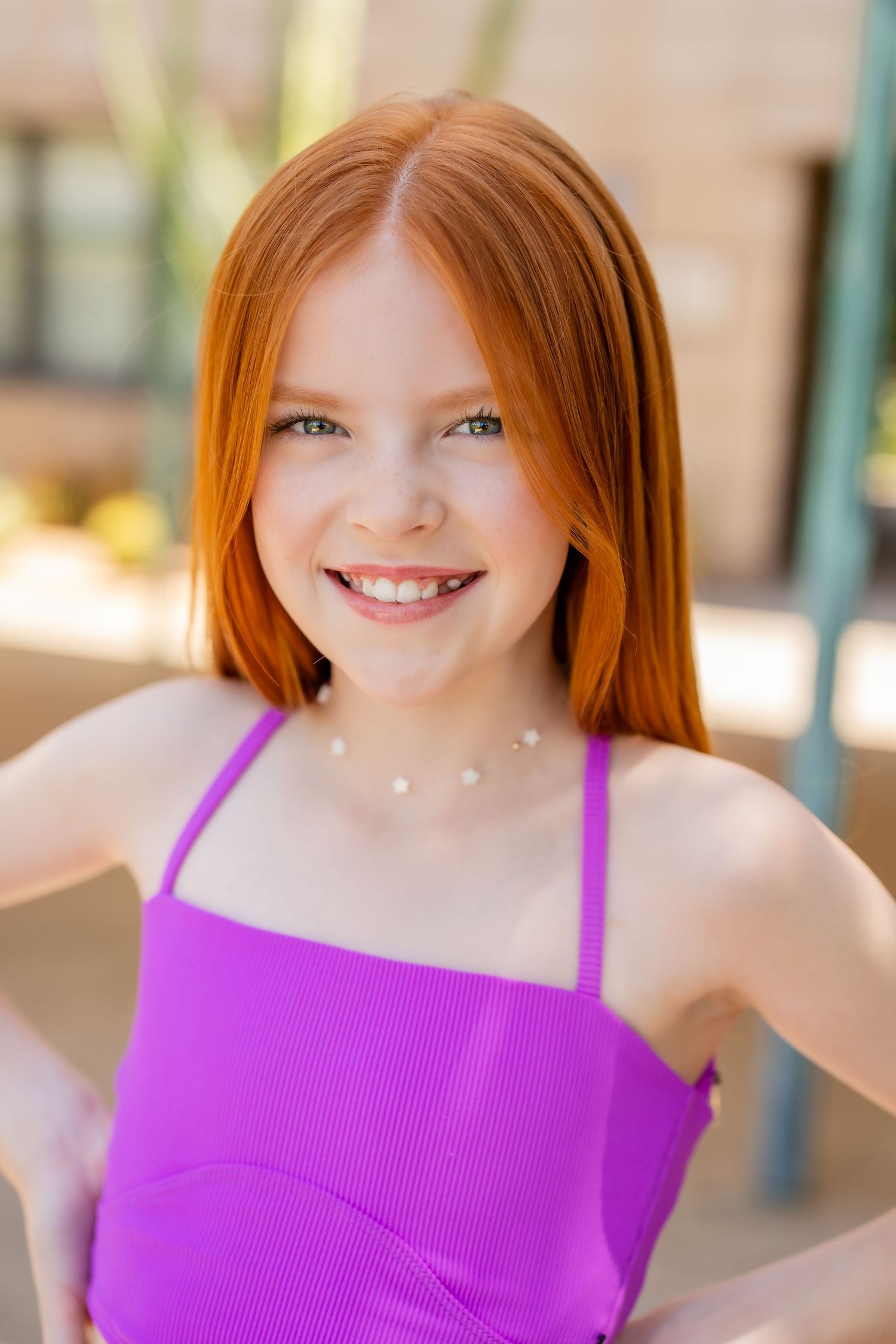 Young person with vibrant red hair wearing a purple tank top smiling at the camera in outdoor lighting.