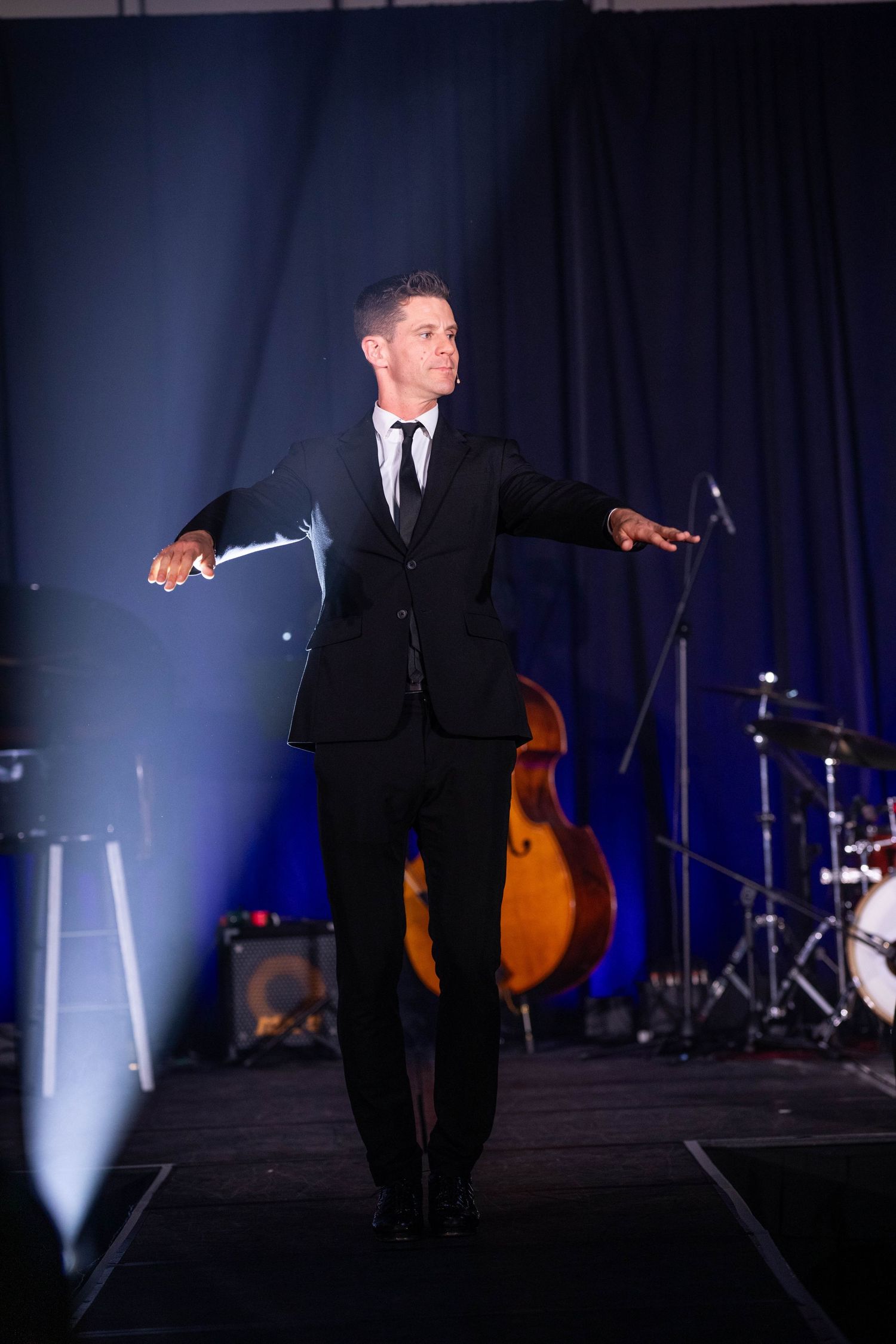 Performer in black suit on stage with musical instruments in background.