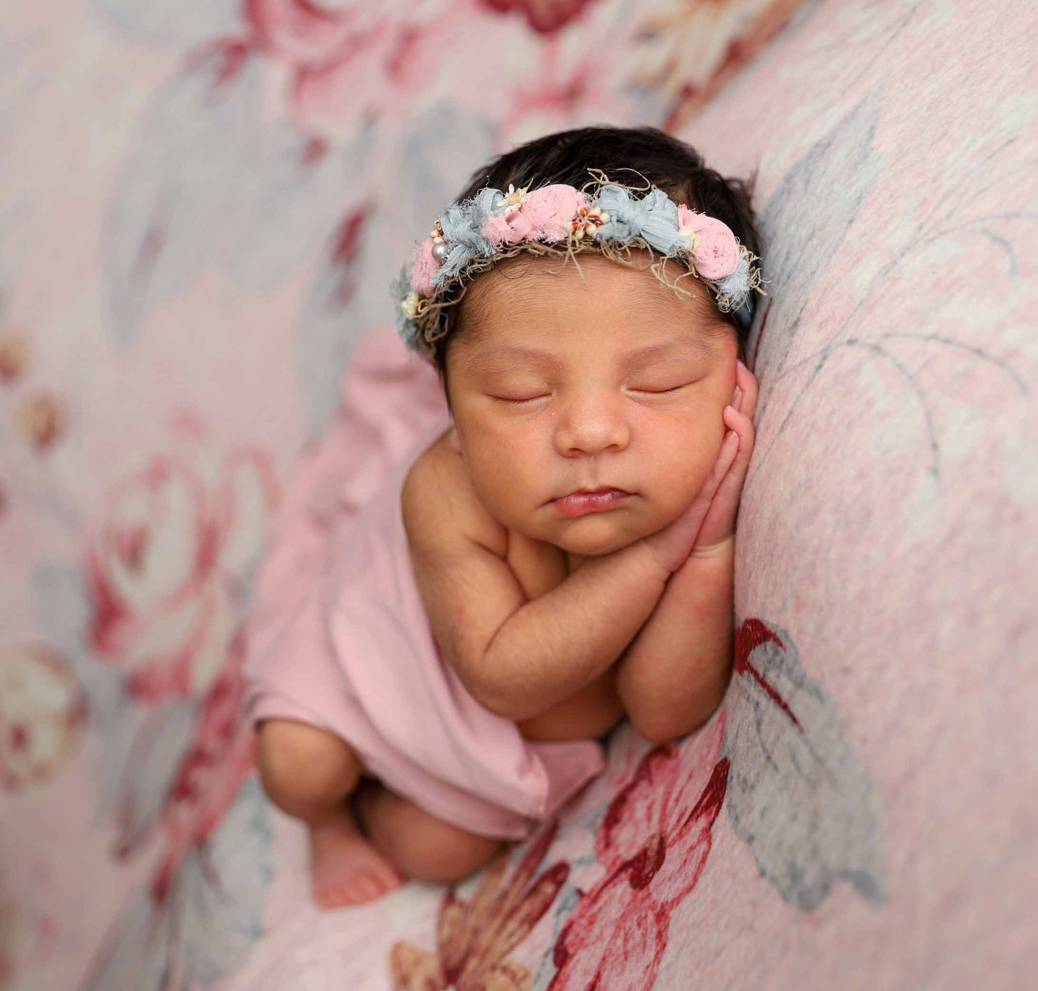 Sleeping newborn baby girl wearing a floral crown headband wrapped in pink on a floral backdrop.