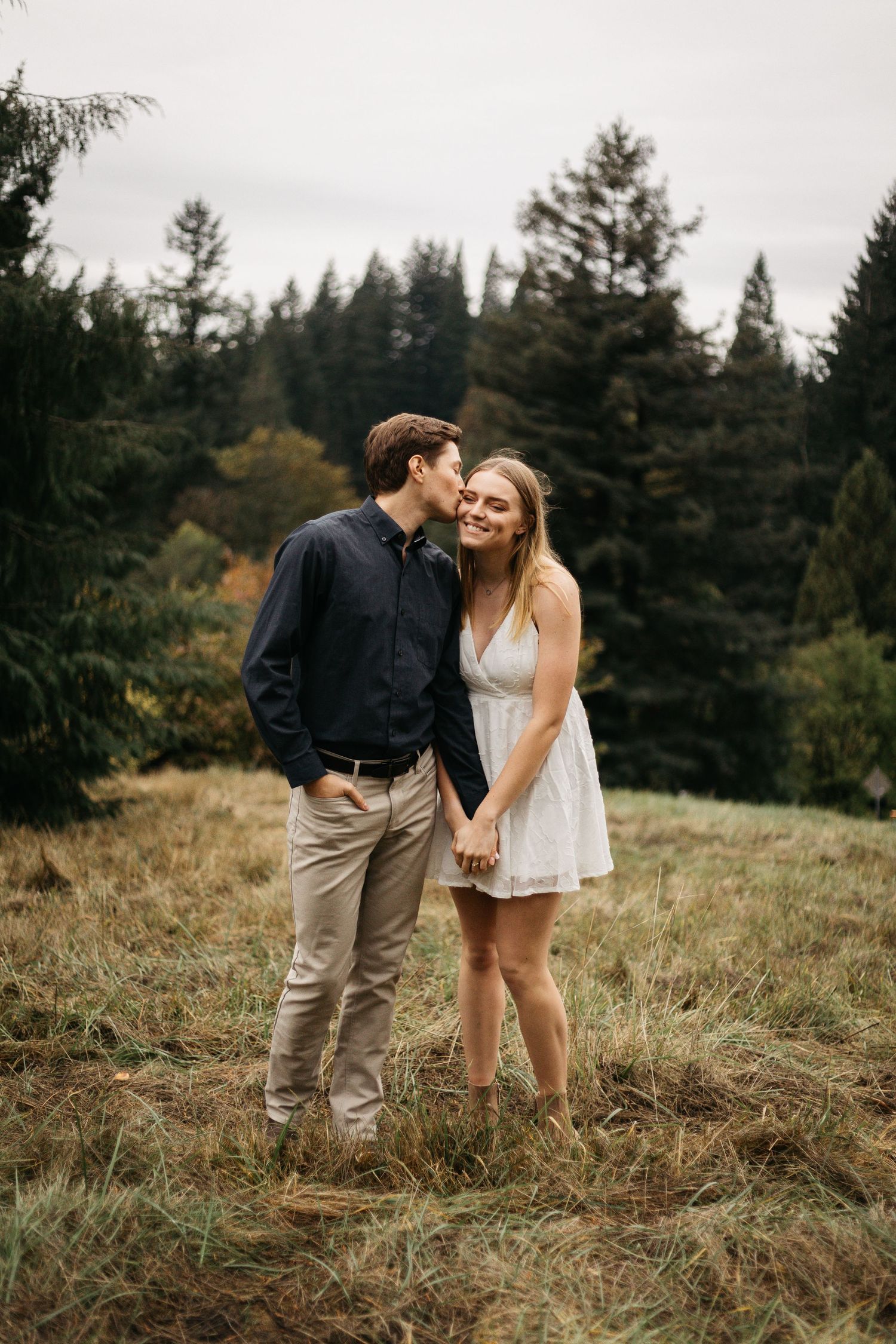 A romantic couple sharing a loving moment on a grassy hillside surrounded by evergreen trees.