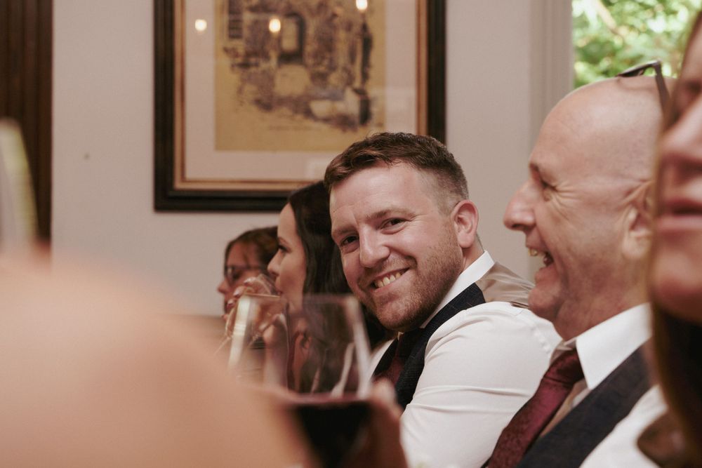 A smiling man in a waistcoat laughs with guests seated at a long wedding table in an elegant dining room.
