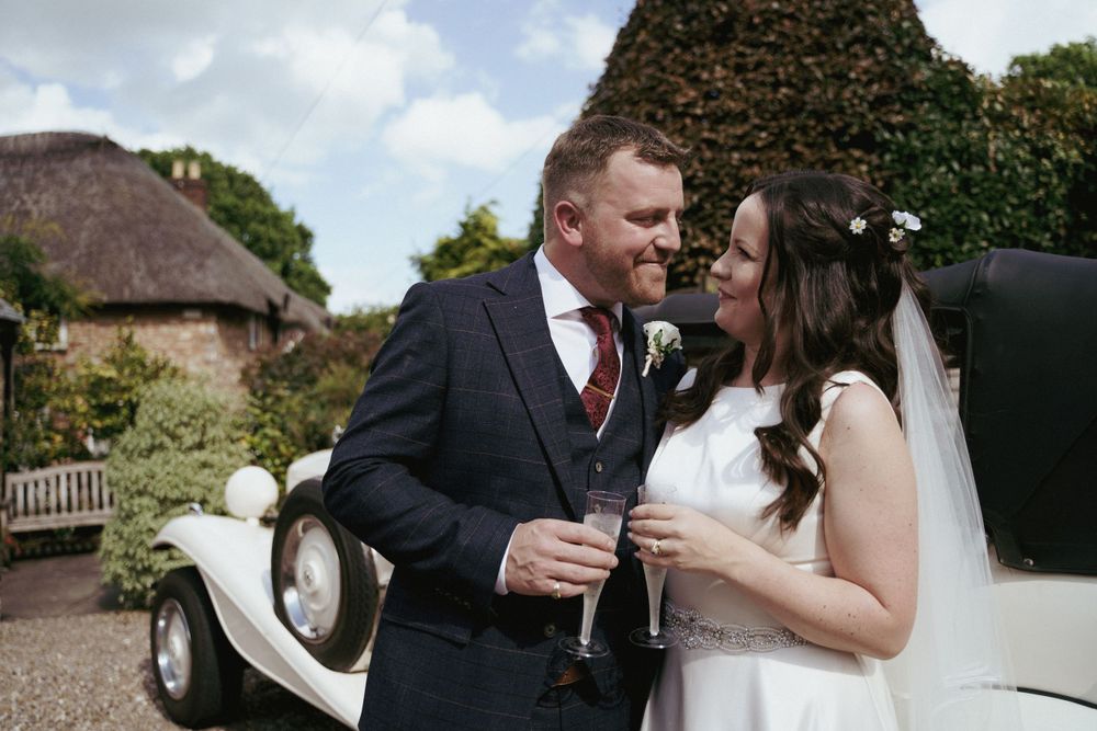 A bride and groom toast with champagne beside a vintage white wedding car in front of a thatched cottage.