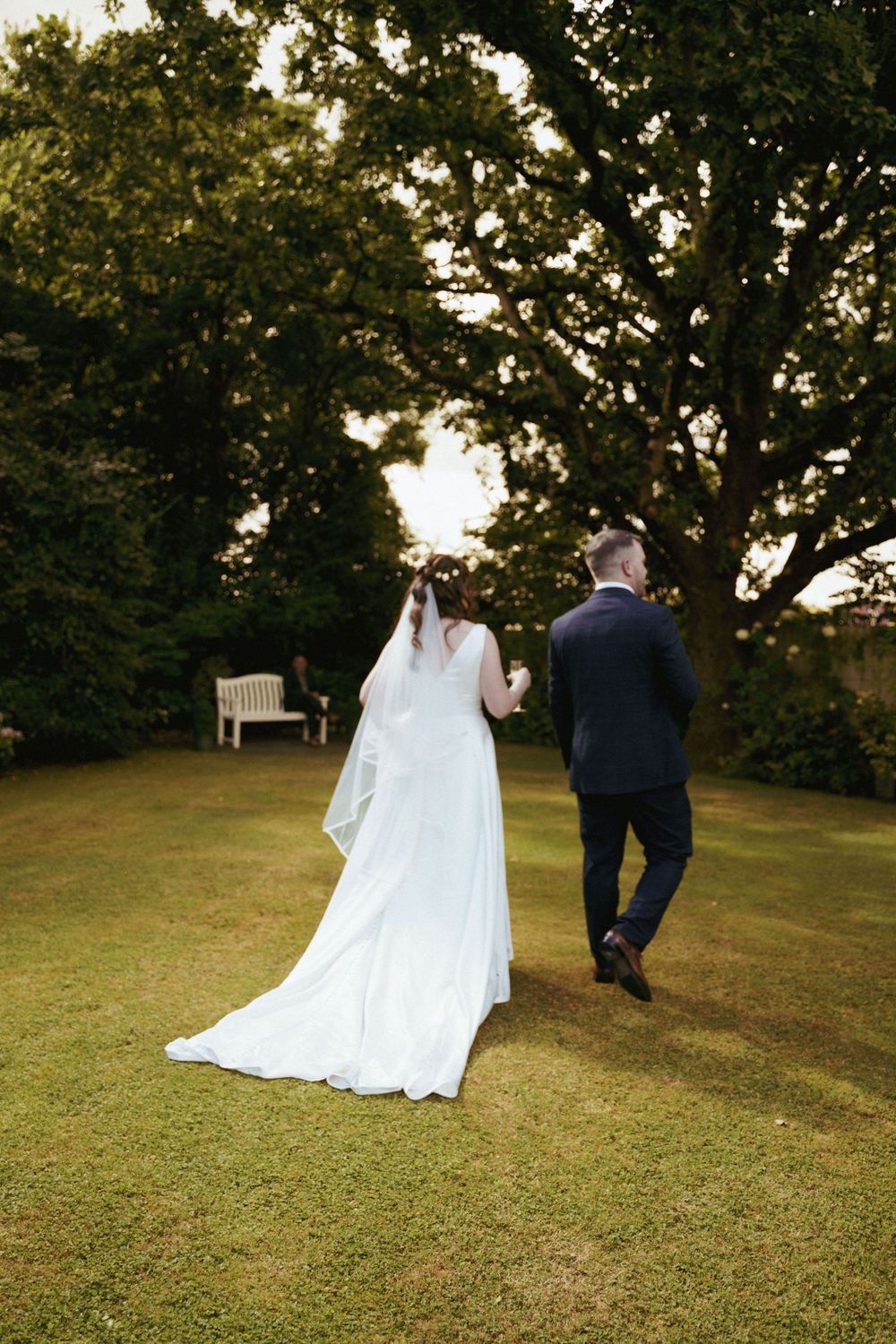 A bride in a long white gown and veil walks hand in hand with her groom across a sunny lawn beneath large trees.
