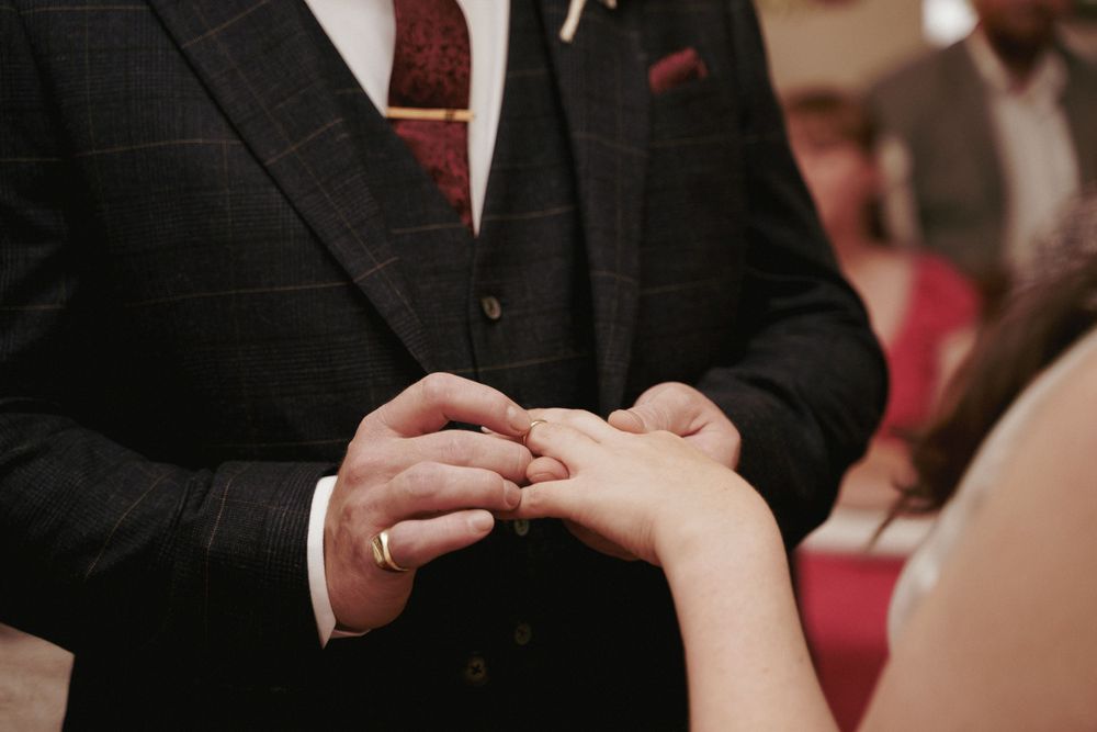 A groom in a dark checked suit places a gold wedding ring on the bride's finger during the ceremony.