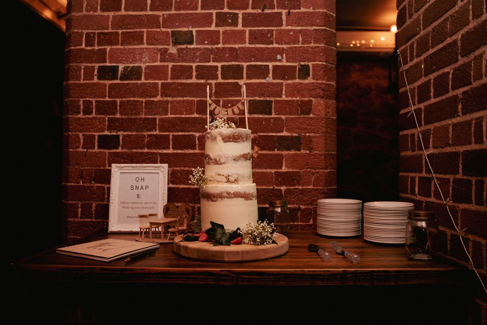 Three-tier semi-naked wedding cake with greenery displayed on a wooden table against an exposed red brick wall.