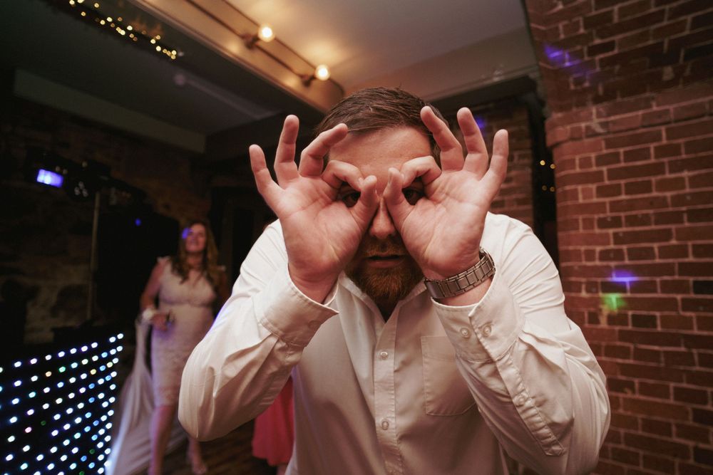 A man in a white shirt playfully makes goggles with his hands over his eyes at a lively indoor party with a brick wall background.