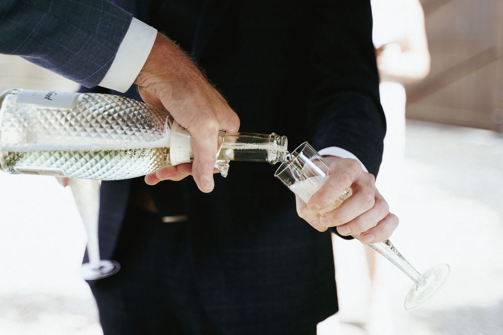 A man in a dark suit pours sparkling wine from a bottle into a champagne flute at a wedding celebration.