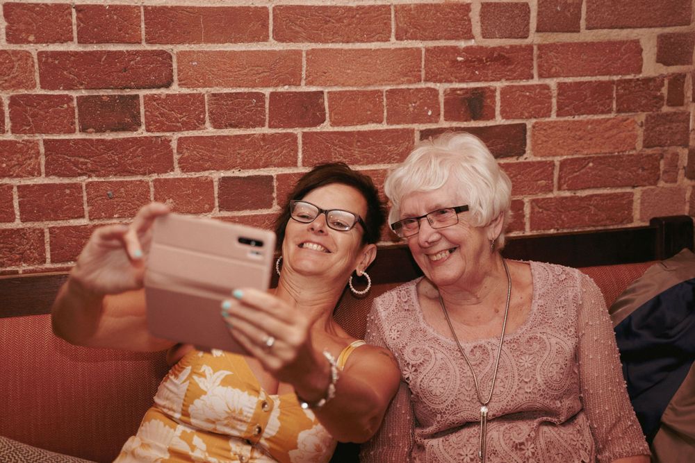 Two smiling women, one with glasses and one with white hair, take a selfie on a phone in front of a rustic red brick wall.