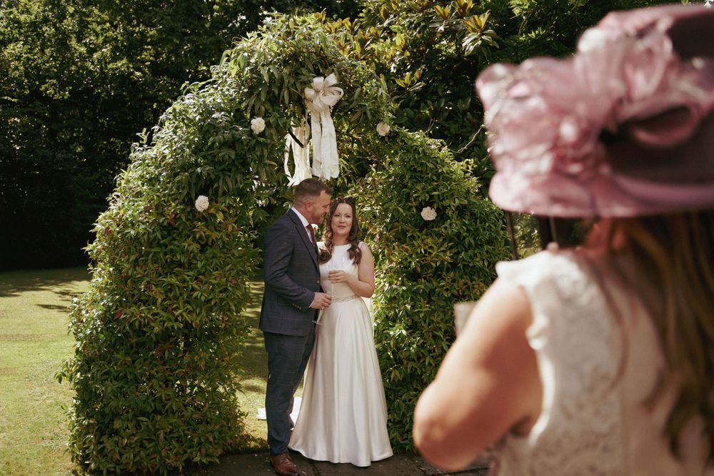 A bride and groom embrace under a green floral garden arch while a guest in a pink fascinator watches.