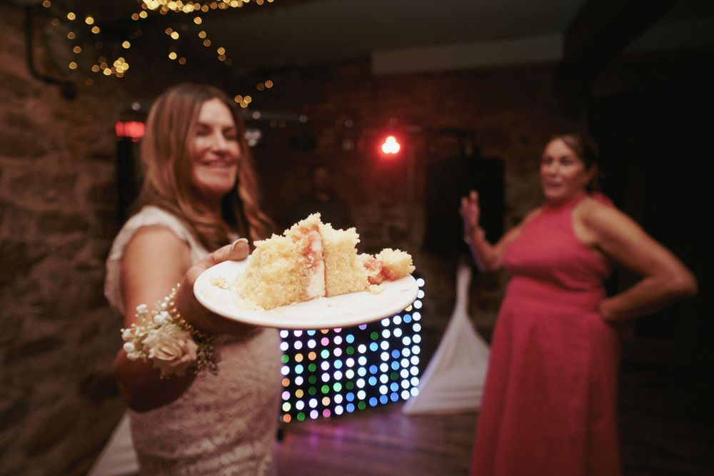 A smiling woman in a lace dress holds out a plate of wedding cake at an evening reception with a colorful light display behind her.