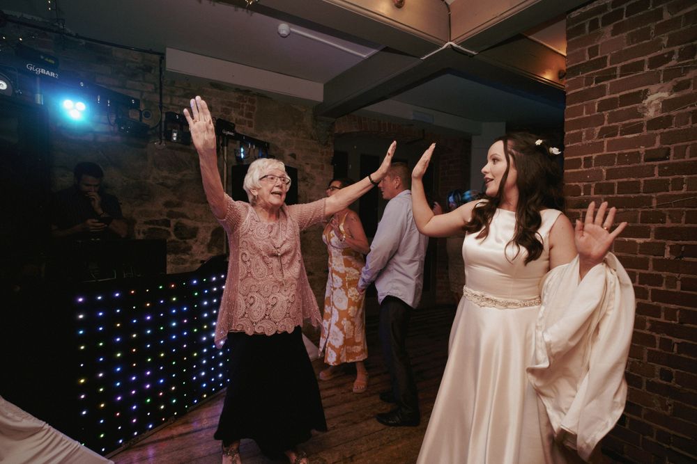 A bride in a white gown and an older woman in pink raise their arms joyfully on the dance floor at a wedding reception.