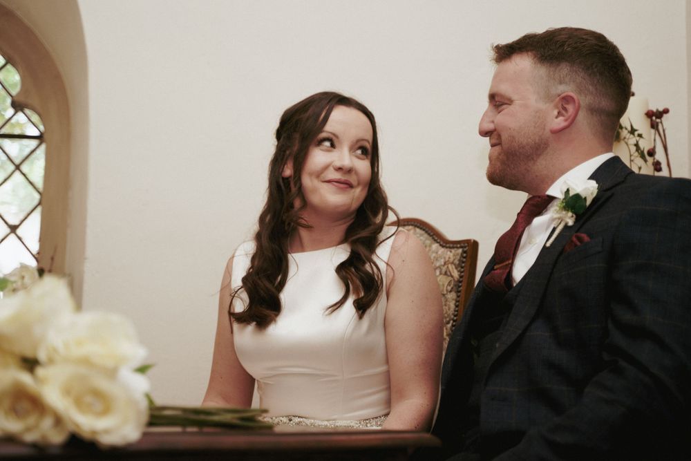 A smiling bride in a white dress gazes at her groom in a dark suit as they sit together at the wedding ceremony table.
