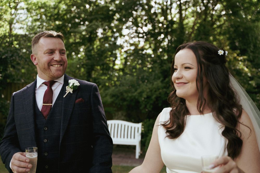 A laughing groom in a tweed suit stands beside his smiling bride in a white dress near a garden bench.
