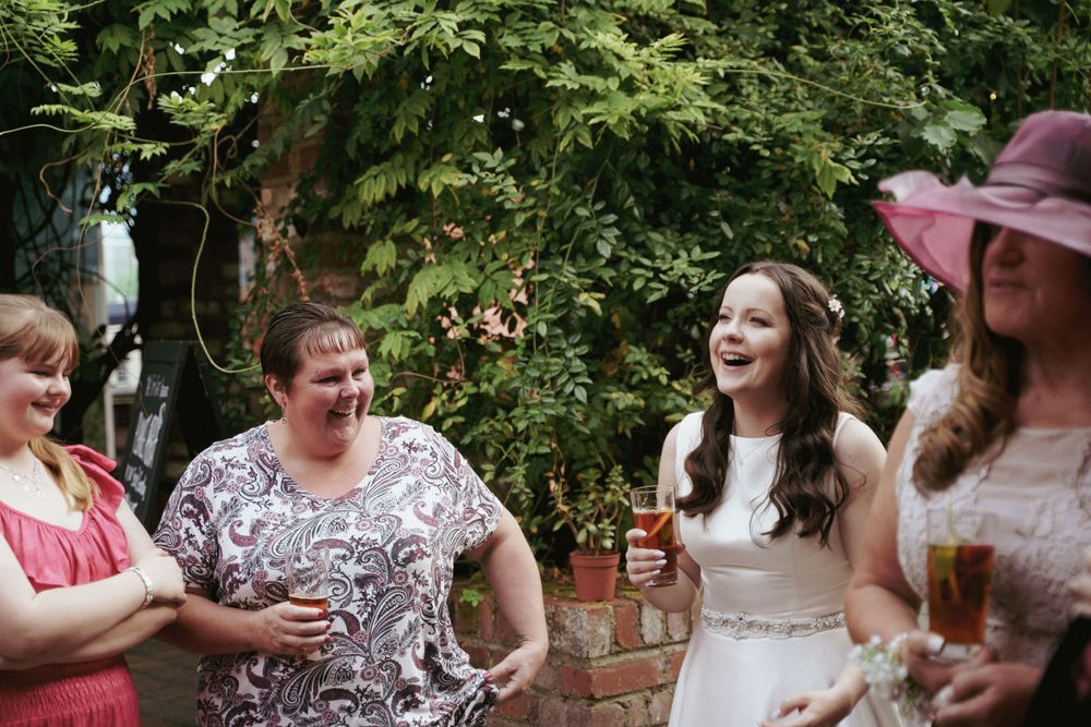 A bride laughs with wedding guests holding drinks in a leafy garden during a summer wedding celebration.