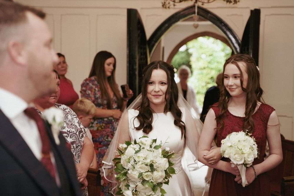 Bride holding a white floral bouquet walks down the aisle beside a bridesmaid in burgundy as guests look on.