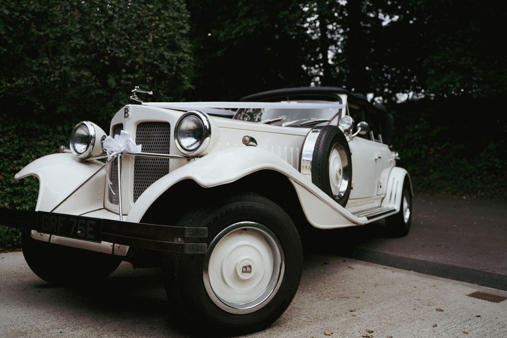 Vintage white wedding car with ribbons parked beneath dark green trees, ready for the bride and groom.