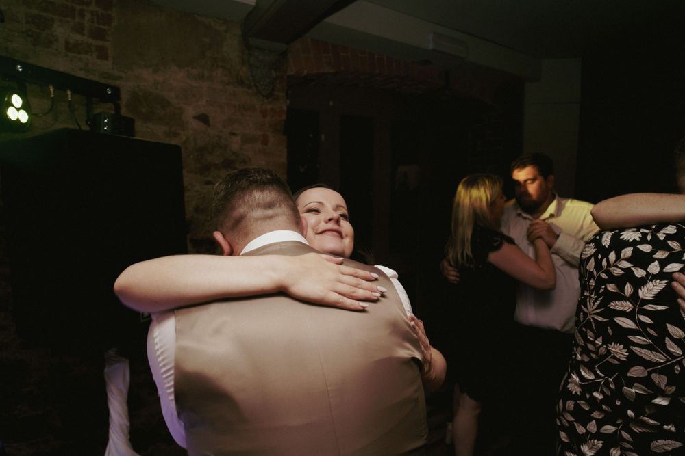 A groom in a waistcoat embraces a smiling bride in a warm hug while guests dance in the dimly lit background.