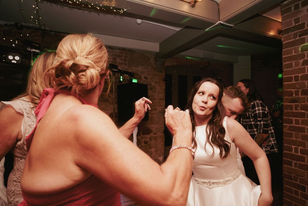 A bride in a white dress dances with wedding guests in a brick-walled reception venue with twinkling lights overhead.