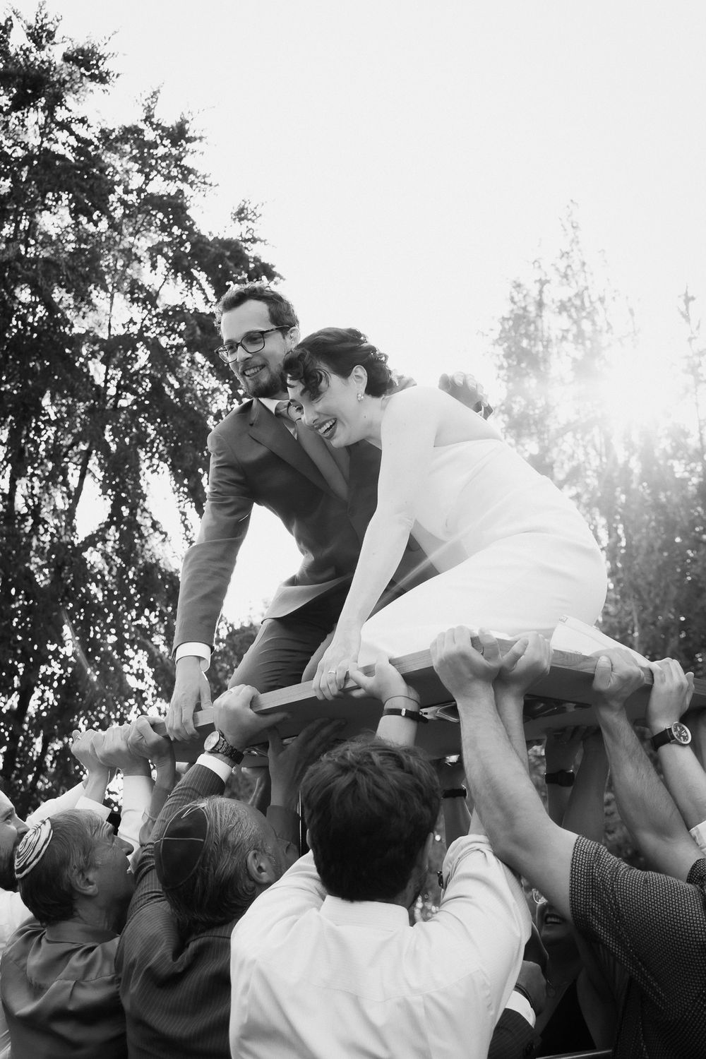 A newly wed couple is being hoisted on a table by family and friends in their jewish ceremony. White dress, grey suit.