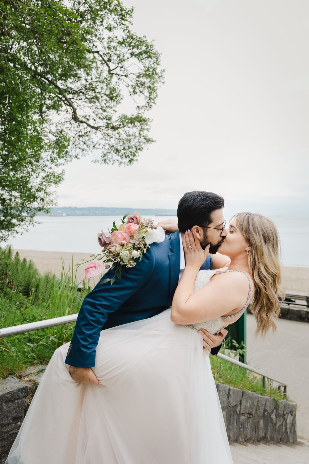 A man dips his new bride, kissing her, with the ocean behind them. She holds her bouquet around his back.