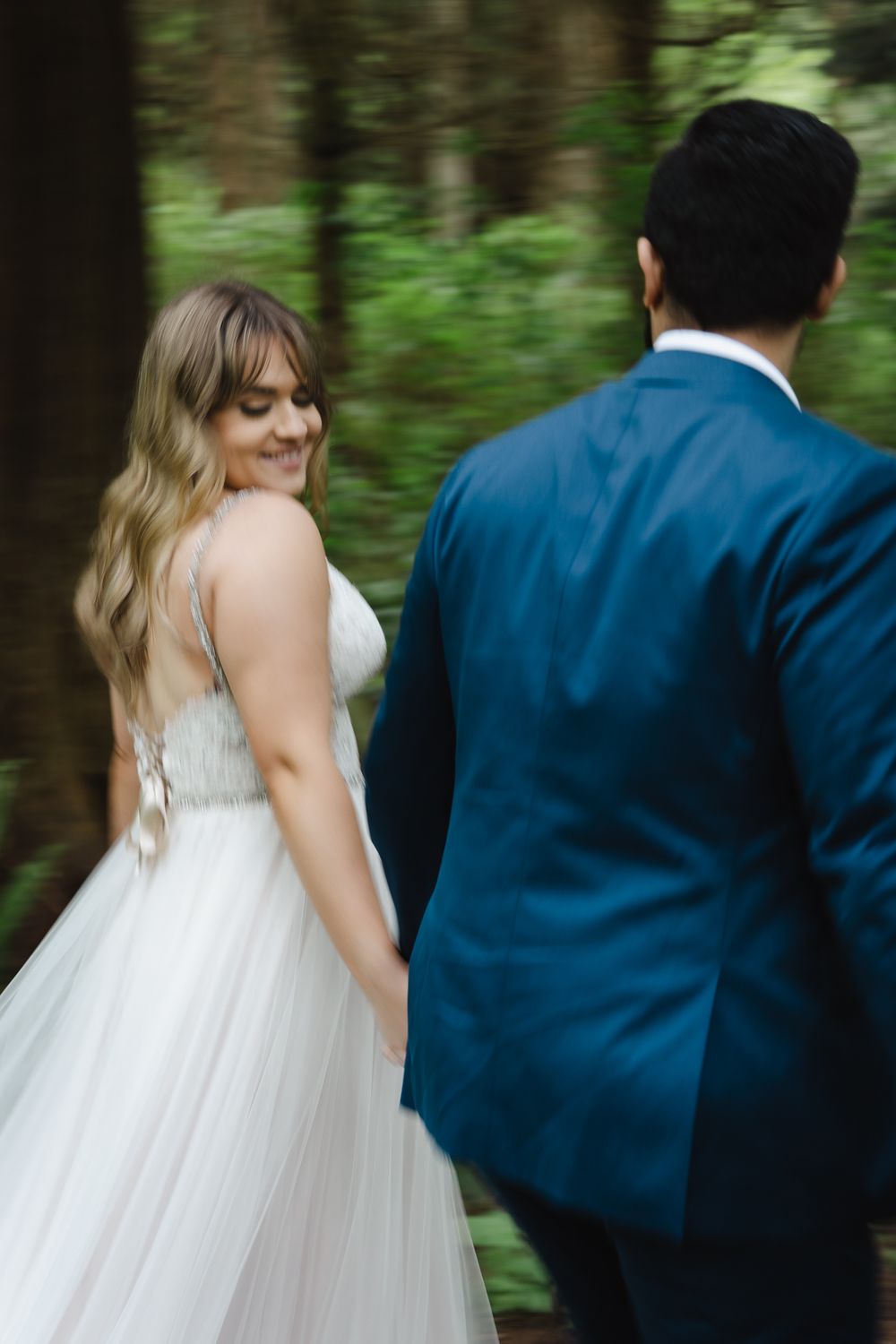 A newly wed couple walks in the forest. The bride looks back at the camera as her husband looks at her. There is motion