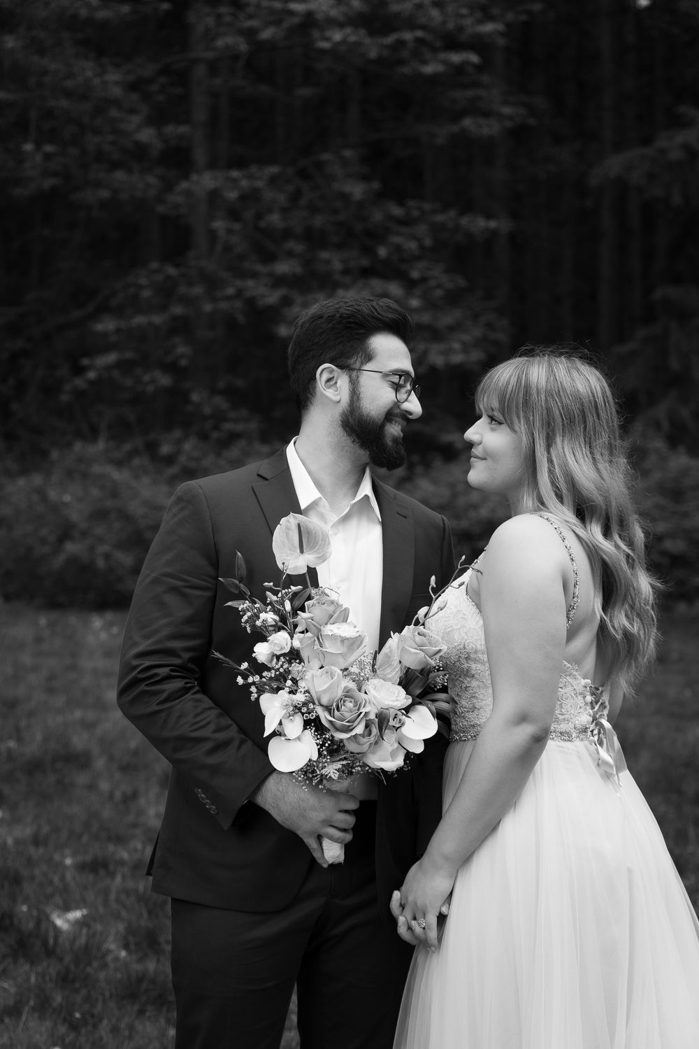 A bride and groom look lovingly at each other while holding hands. He holds her bouquet at his chest.