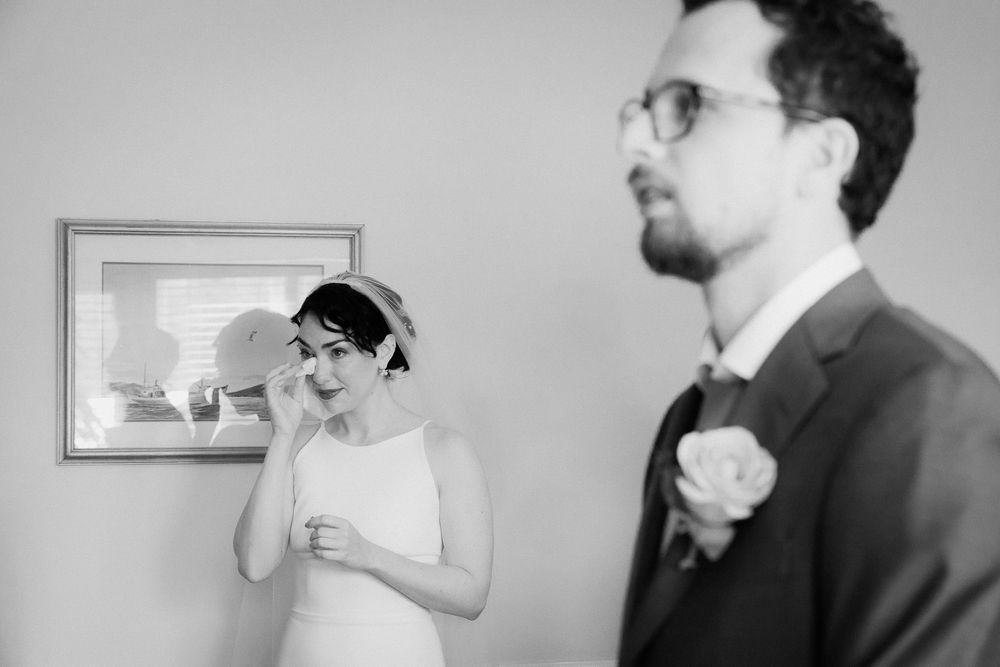 A bride wipes her tears after an emotional ceremony. Her groom stands in front of her, blurred out to the camera.