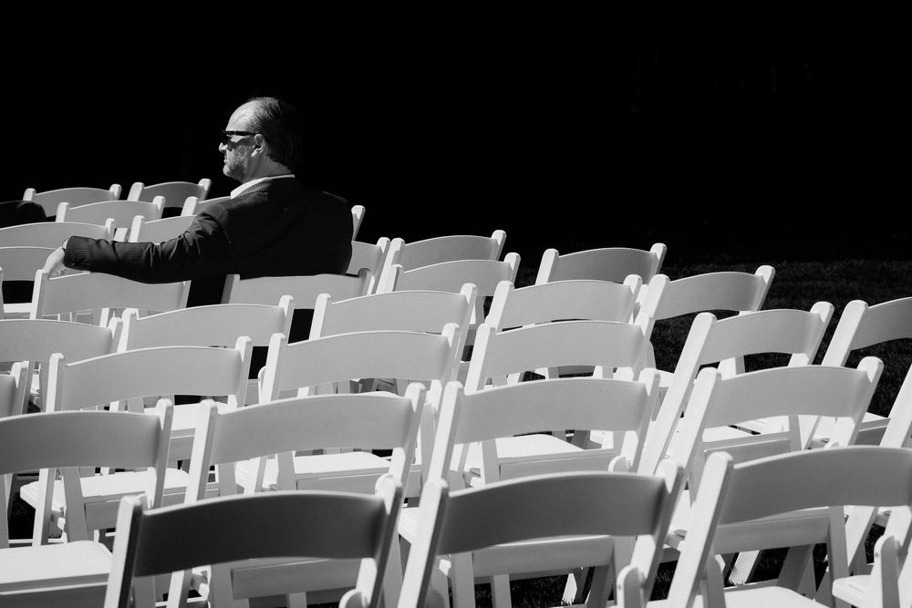 A man sits alone in a sea of white chairs, awaiting the beginning of a wedding ceremony