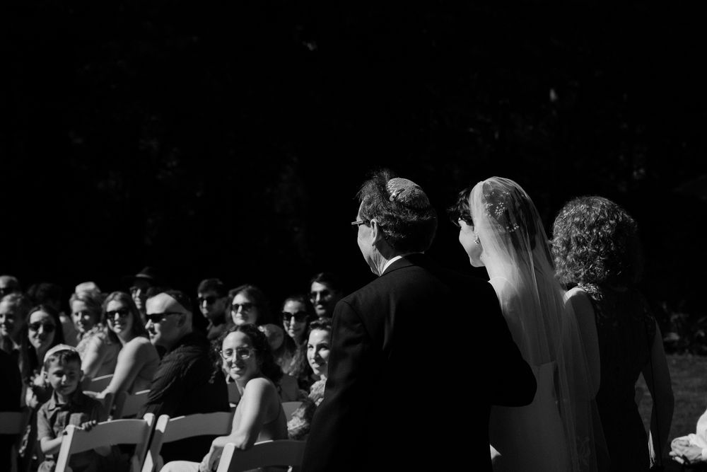 A jewish bride walks down the aisle with both her mother and father beside her. Her veil flows