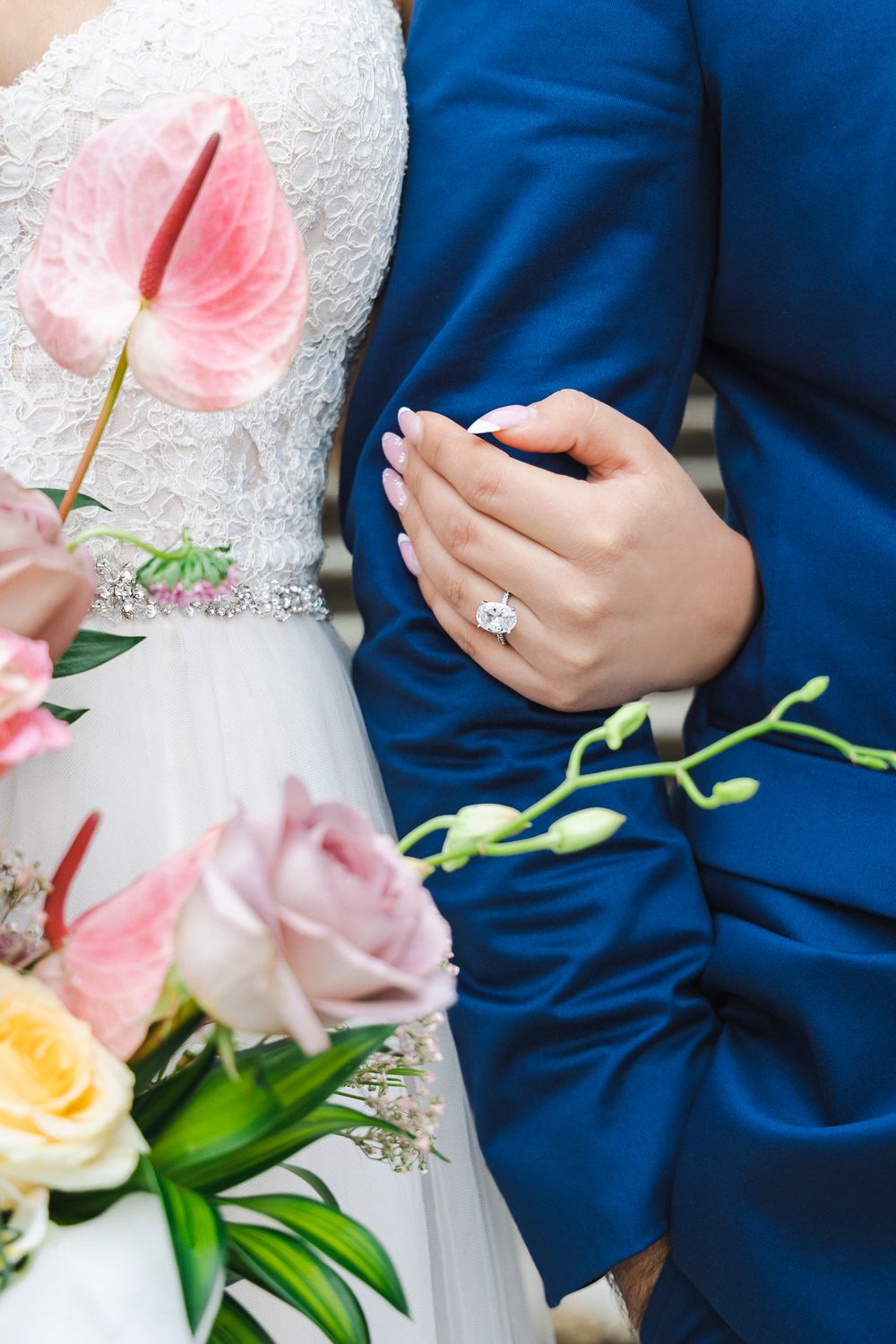 A bride holds her groom's arm, her ring faces the camera, her bouquet is beside her. Close shot.