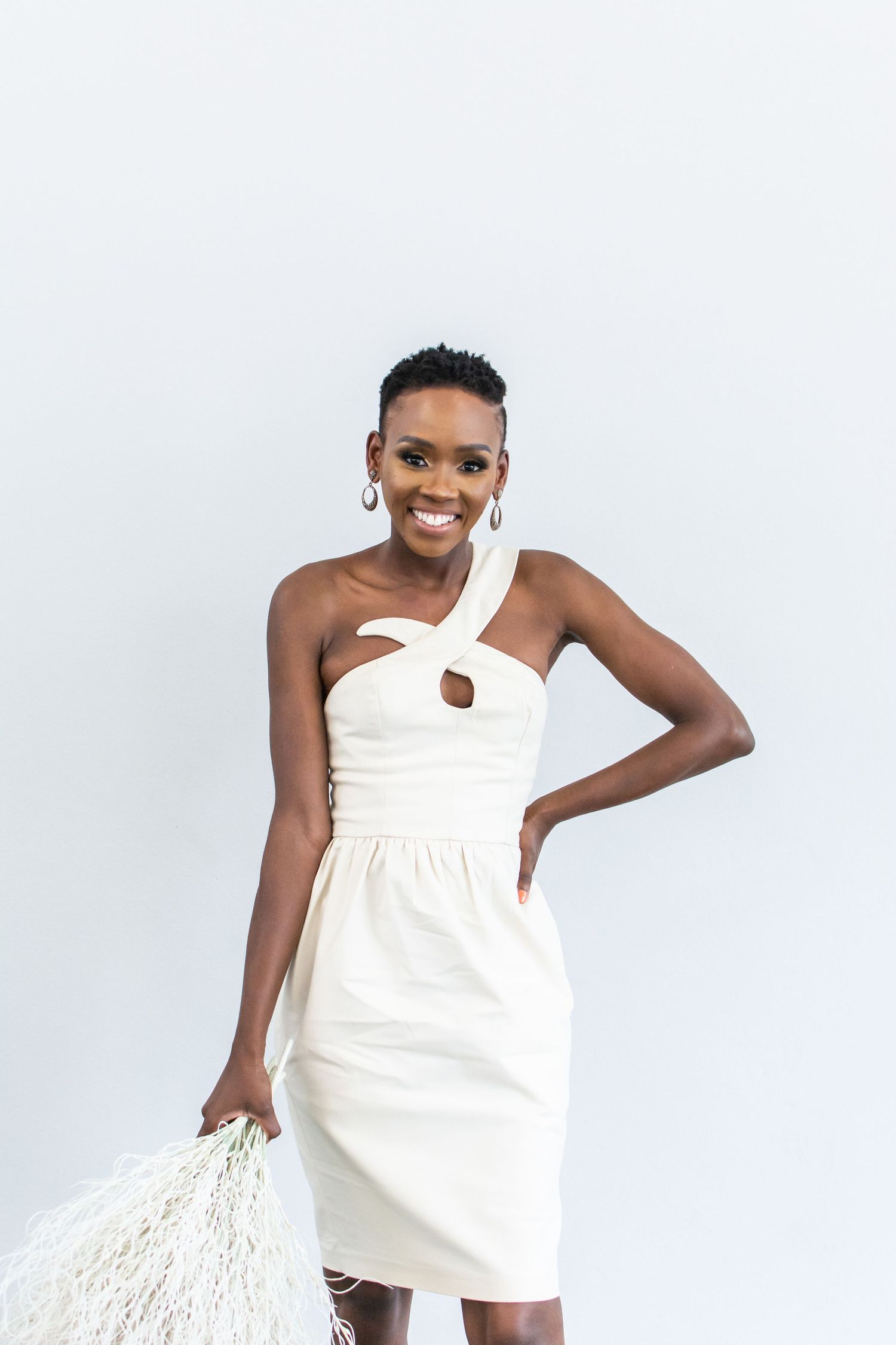 Elegant white one-shoulder dress photo with macrame bag against white background.
