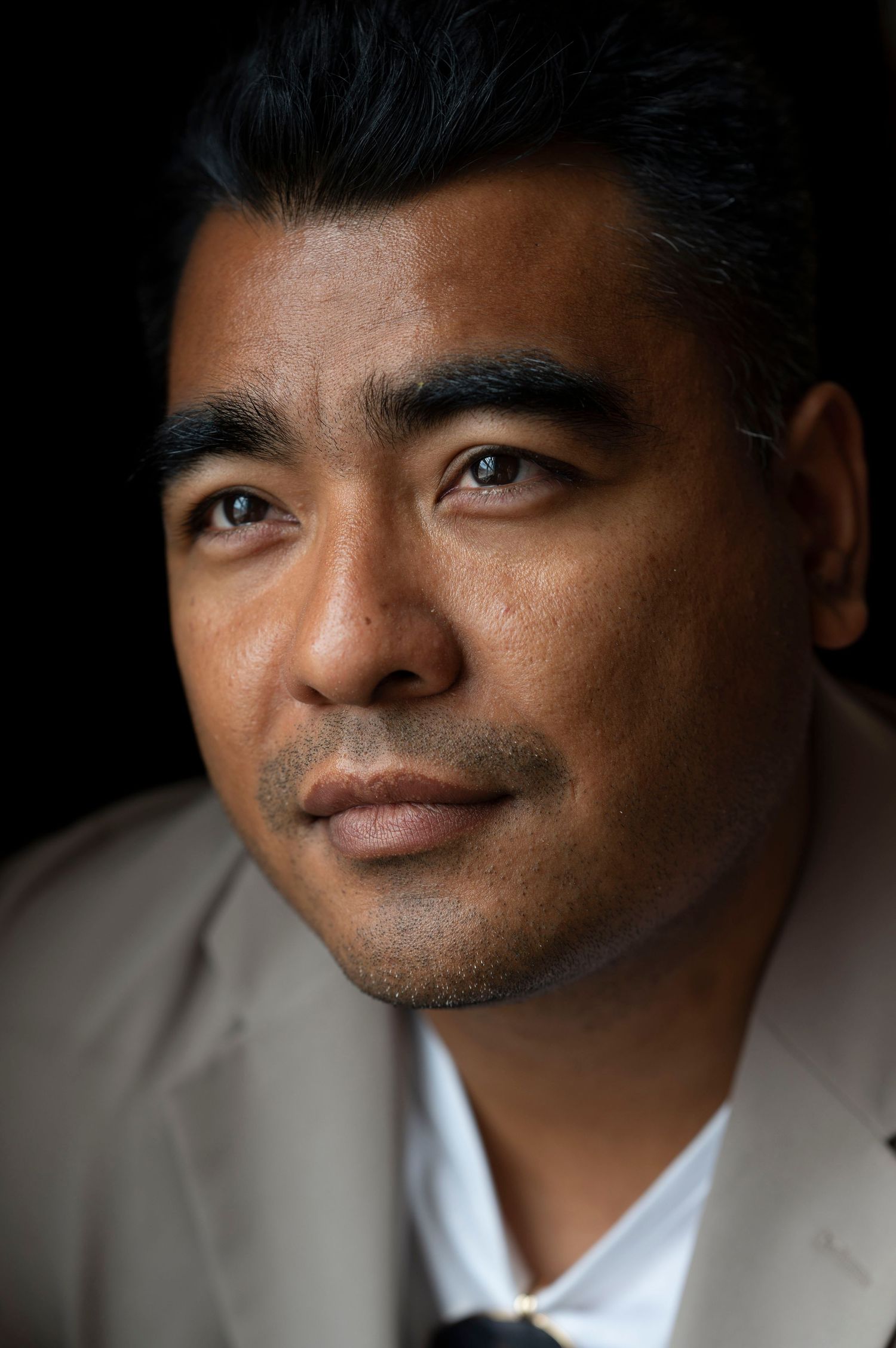 Close-up portrait of a man in a beige blazer against a dark background.