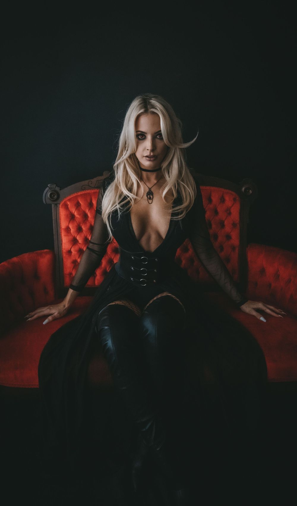 Blonde woman reading a red book titled 'Let Them Fear Her' seated on dark floor before ornate red velvet sofa.