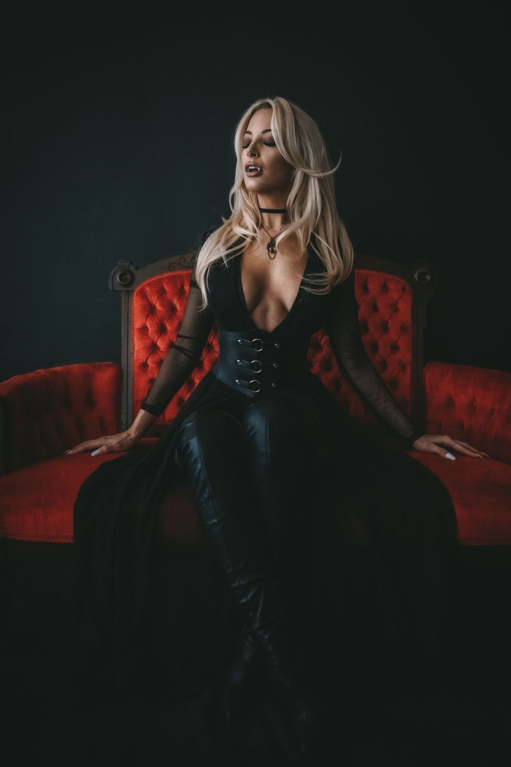 Blonde woman reading a red book titled 'Let Them Fear Her' seated on dark floor before ornate red velvet sofa.