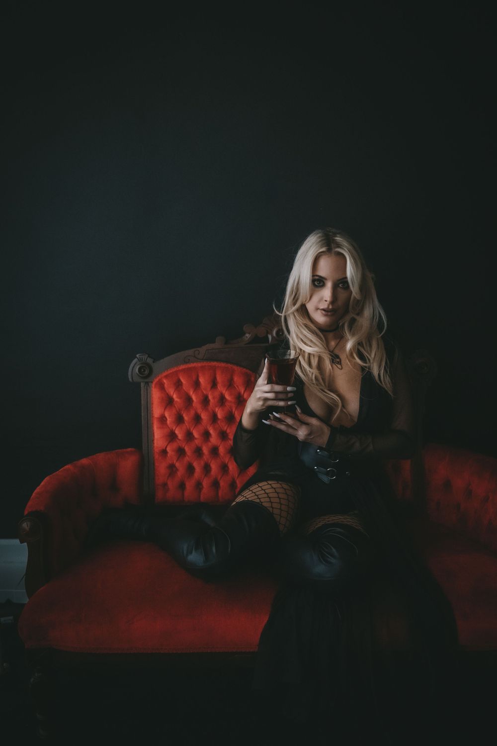 Blonde woman reading a red book titled 'Let Them Fear Her' seated on dark floor before ornate red velvet sofa.