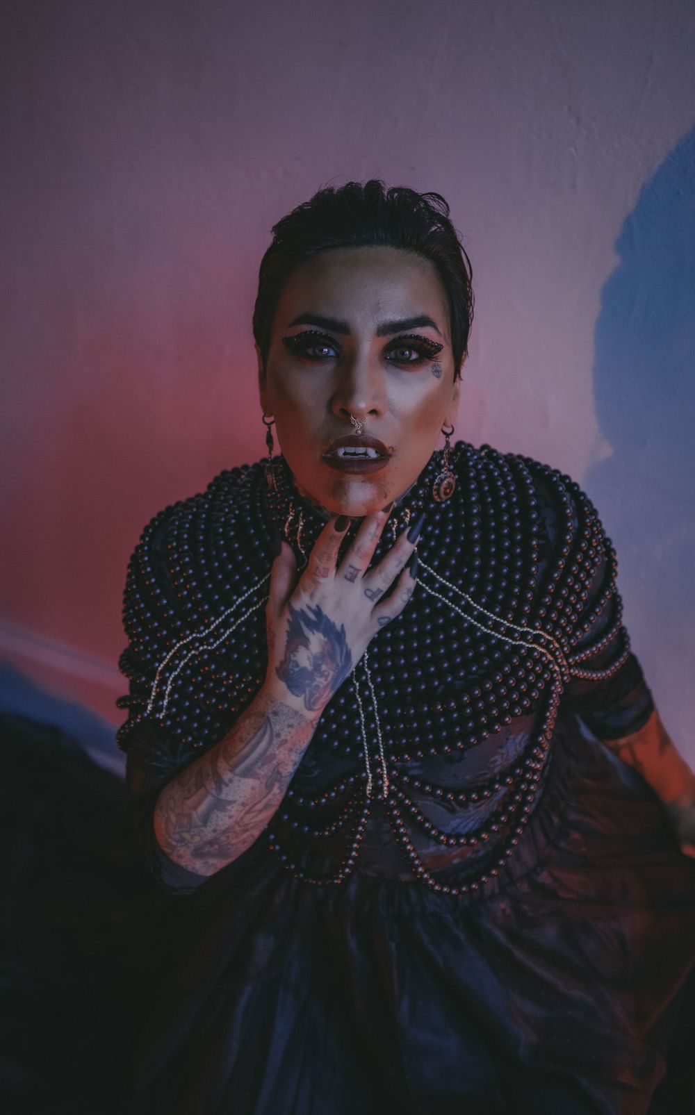 Tattooed woman in black pearl-adorned gown poses dramatically against a pink-lit wall in gothic fashion photography.