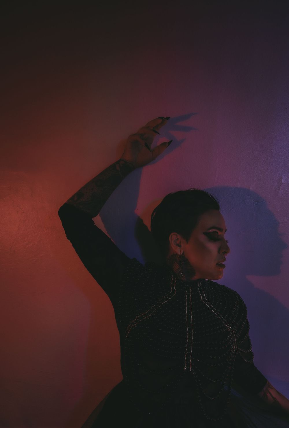 Tattooed woman in black pearl-adorned gown poses dramatically against a pink-lit wall in gothic fashion photography.