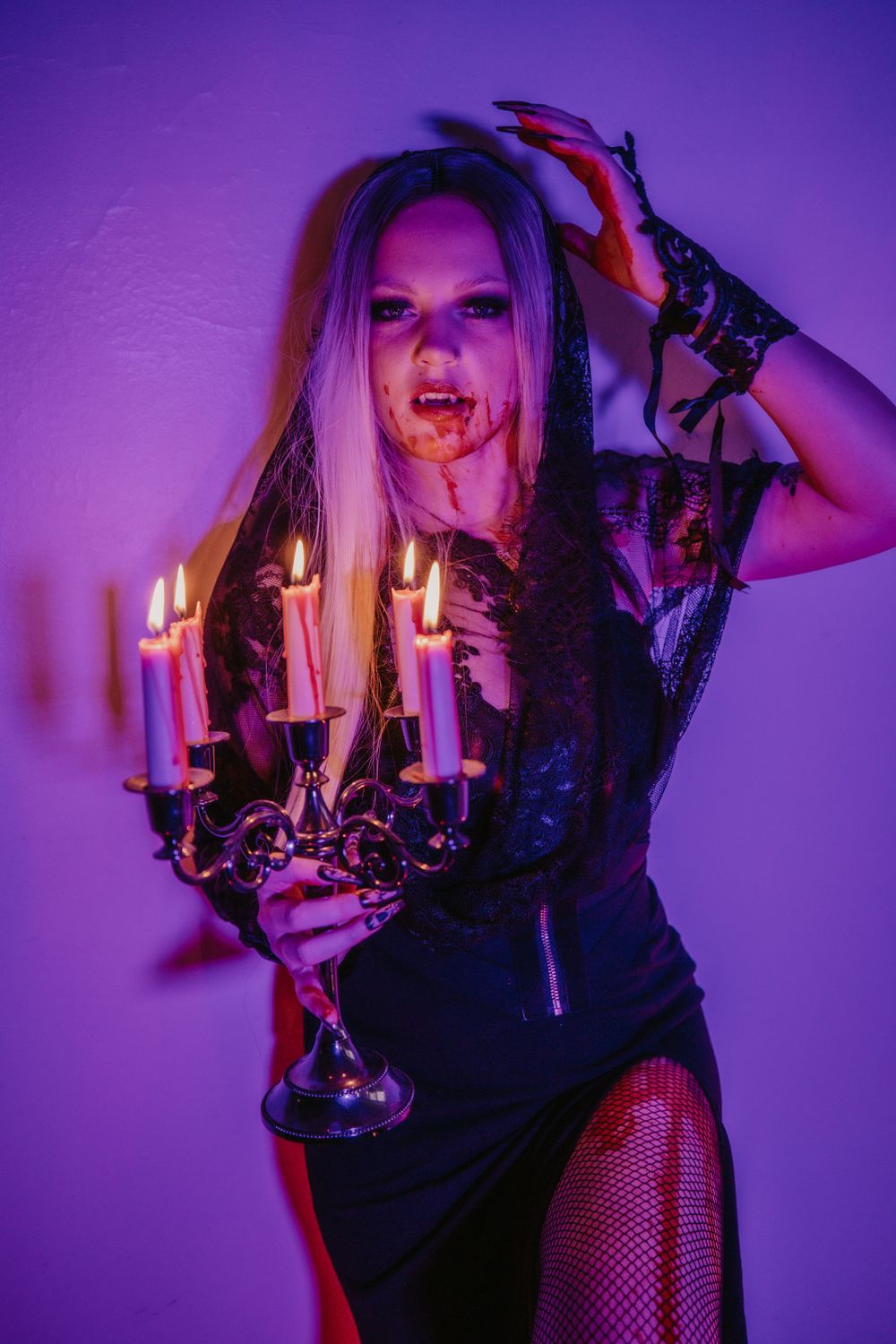 A gothic woman in a black gown holds a candelabra with lit candles, wearing dramatic makeup and fishnet stockings.
