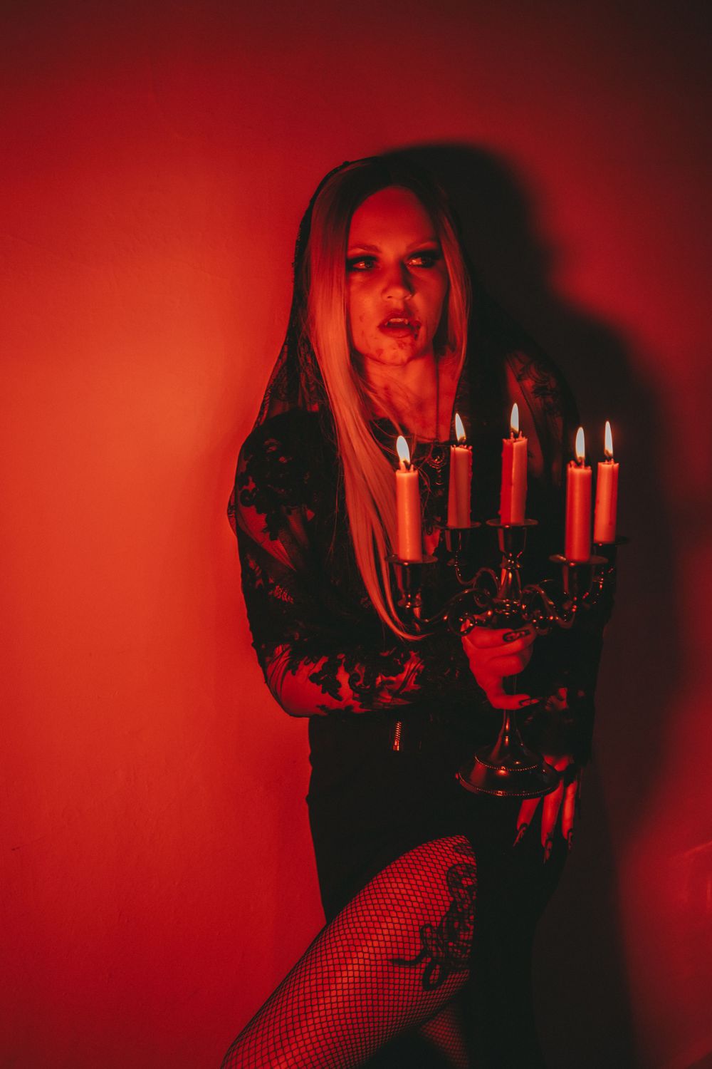 A gothic woman in a black gown holds a candelabra with lit candles, wearing dramatic makeup and fishnet stockings.