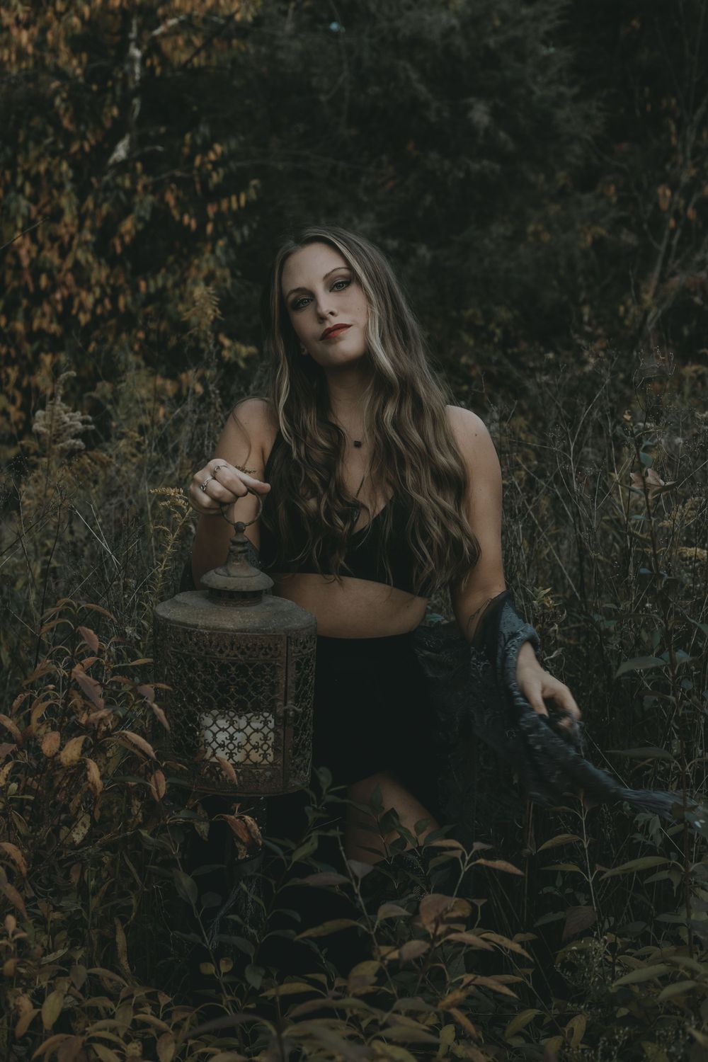 Woman with wavy hair posing with a vintage lantern among autumn forest plants.