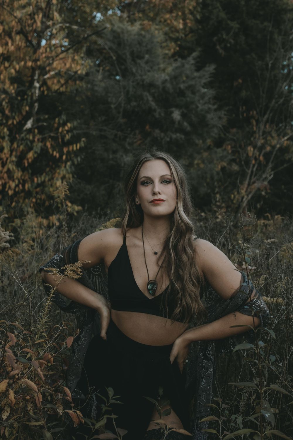 Woman in black outfit sitting among autumn foliage in a moody forest photography session.