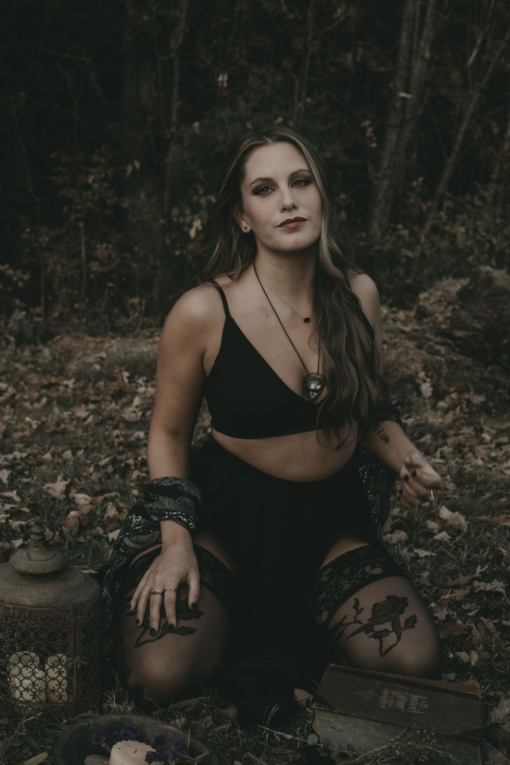 Woman in black crop top and skirt kneeling outdoors with lanterns in dark forest setting.