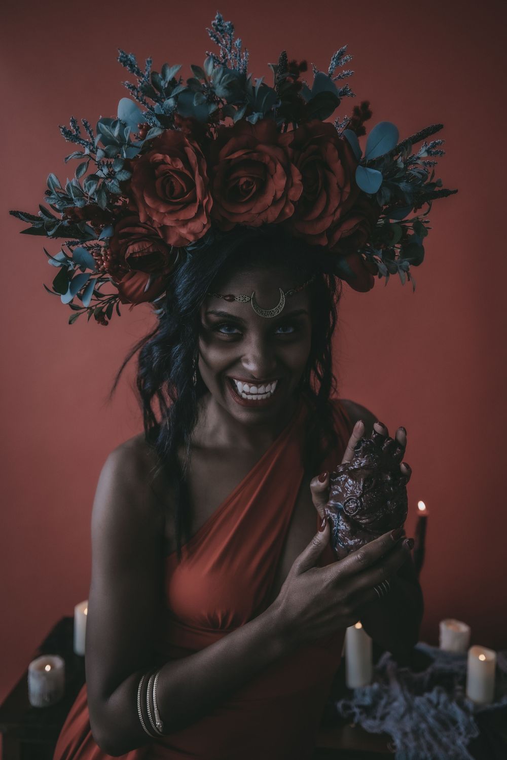 Woman in orange dress and floral crown poses by candlelit altar in moody red room.