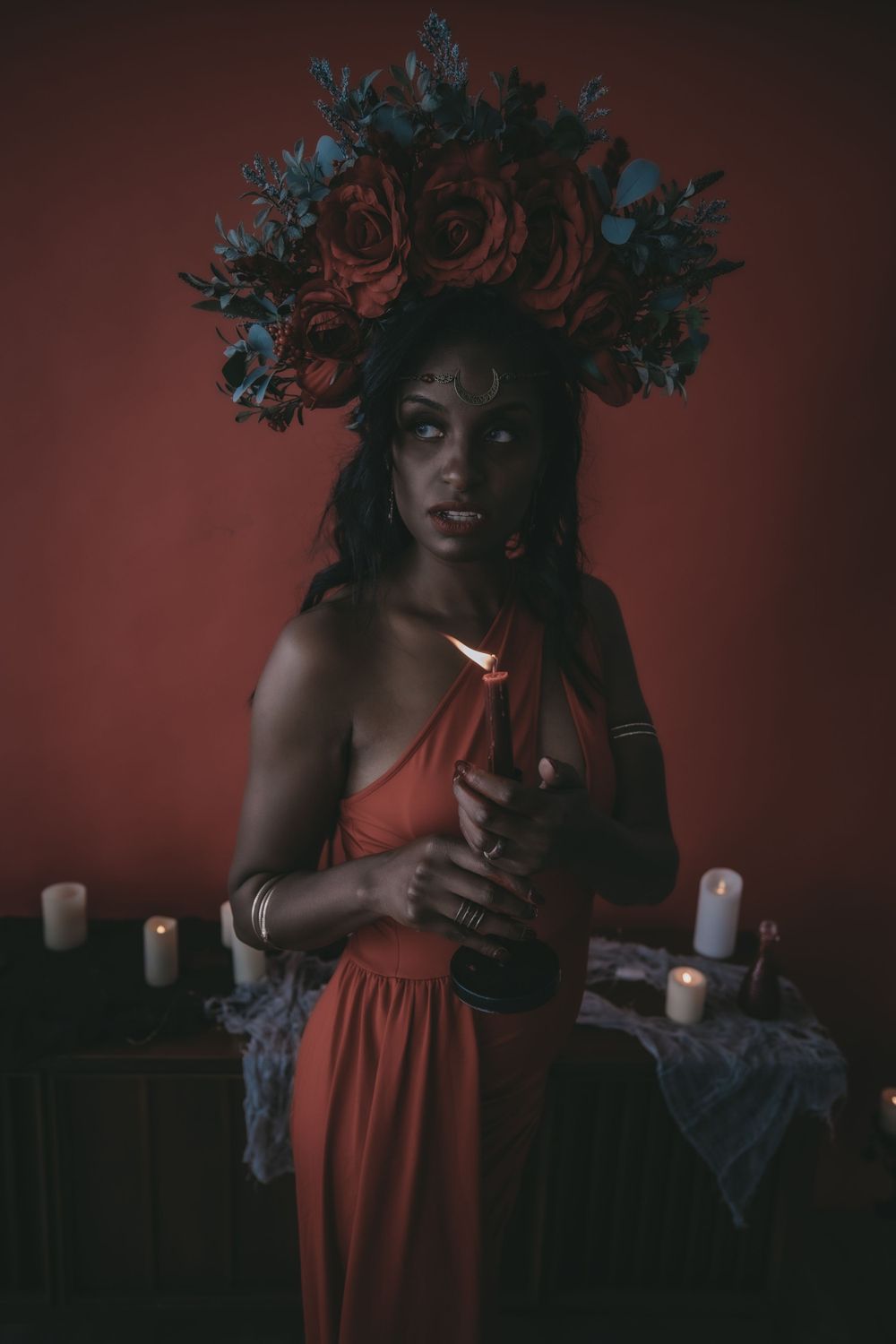Woman in red dress and floral crown holds candle surrounded by lit candles against a dark red background.