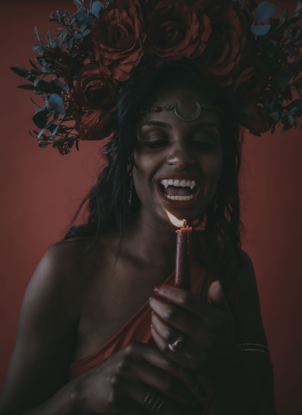 A joyful woman wearing a floral crown of red roses holds a lit candle, laughing against a deep red background.