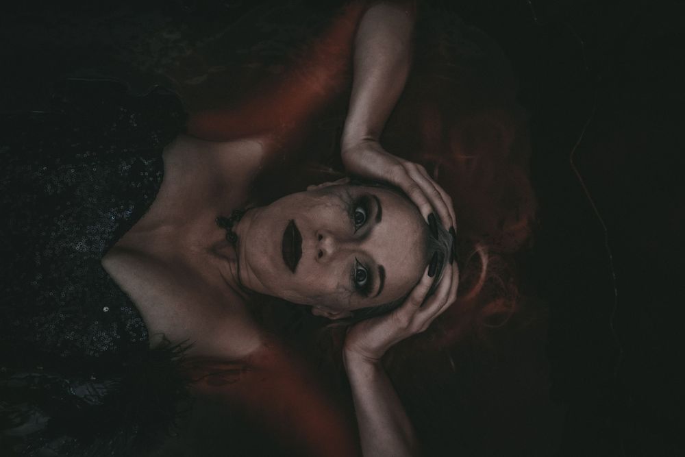 A dramatic vampire woman with red hair, fangs, dark nails, and gothic makeup poses in dark atmospheric lighting.