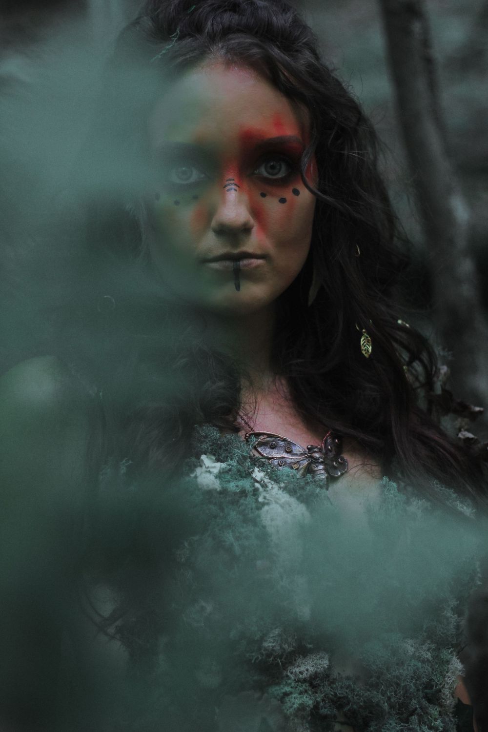 Woman with dramatic face paint and dark makeup peering through green forest foliage.