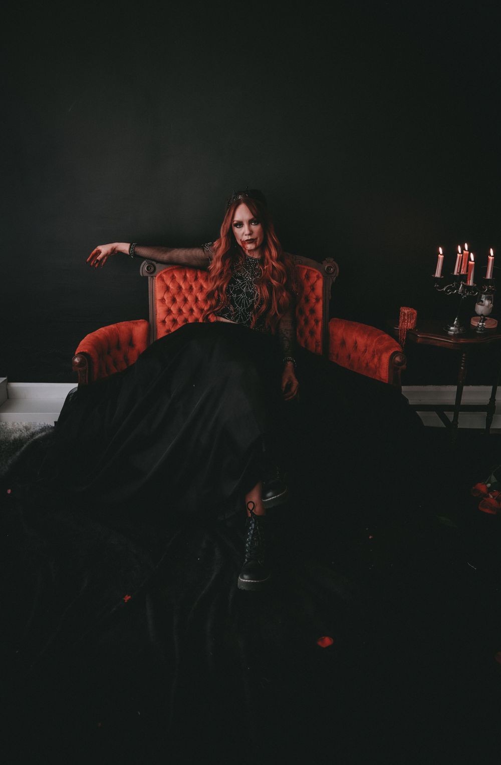 Woman in black gown seated on red velvet throne chair with candles in dark dramatic setting.