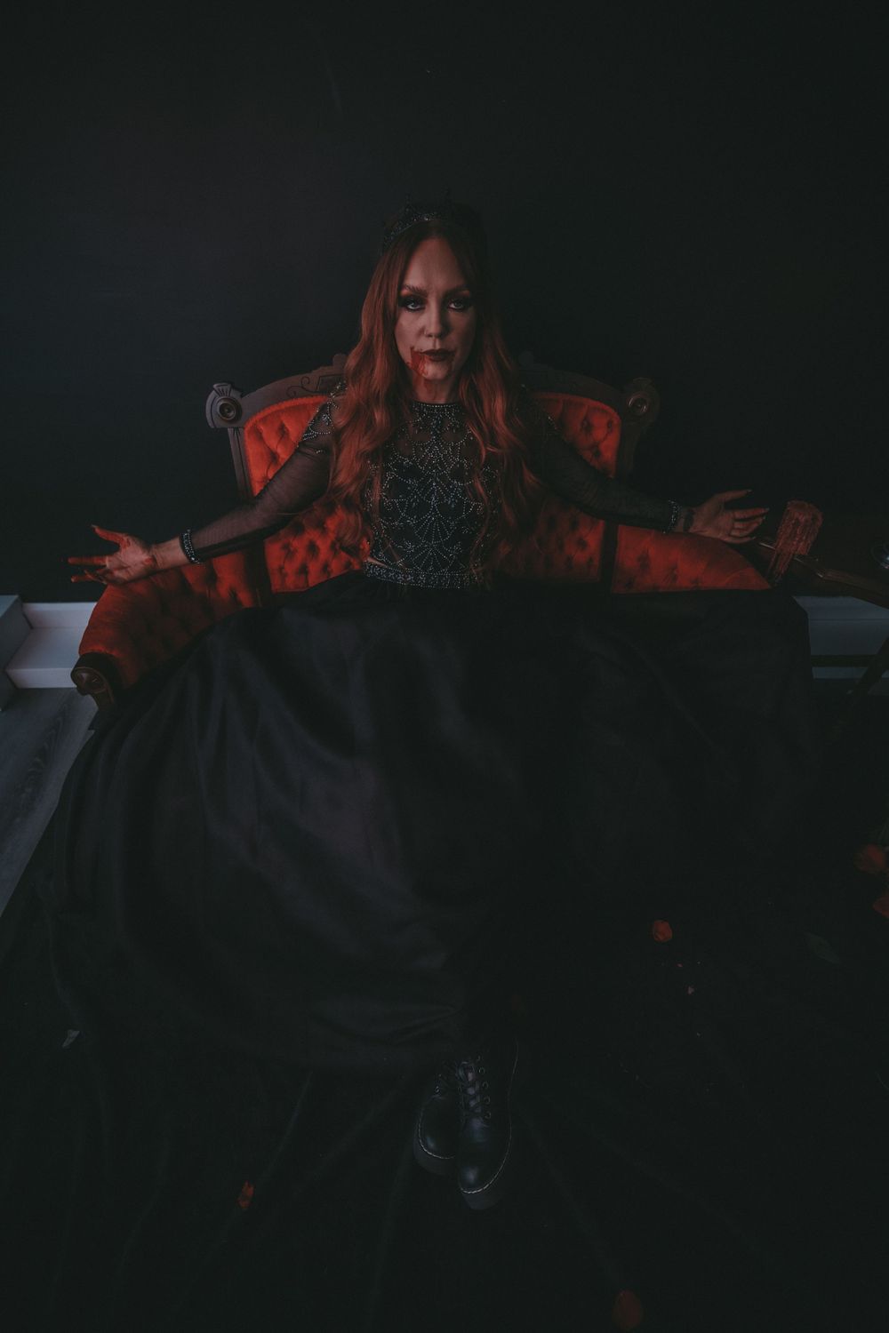 Woman in black gothic gown spreading skirt dramatically on red velvet throne in dark studio.