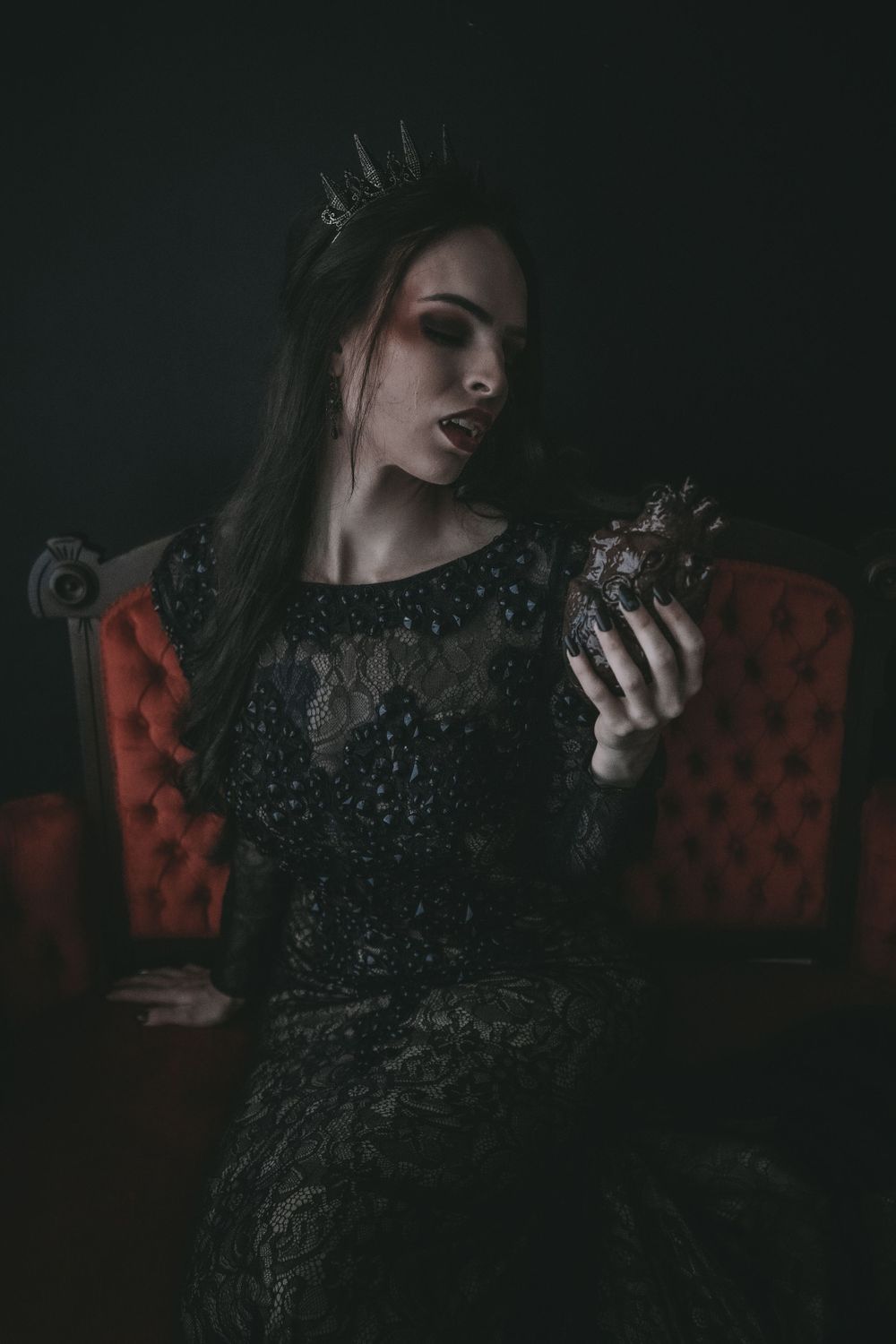 A dark queen in black lace dress and crown sits on red throne holding mysterious orb in moody gothic portrait.